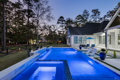 Luxury modern home with illuminated blue swimming pool and hot tub at dusk, surrounded by trees and manicured lawn