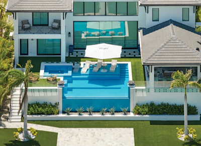 Aerial view of luxury beachfront home with infinity pool, manicured lawn, and modern white architecture overlooking ocean