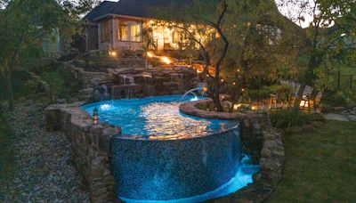 Illuminated backyard pool at dusk with stone features, mature trees, and lit house in background