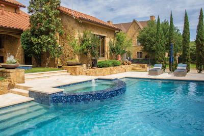 Luxury stone mansion with resort-style swimming pool, spa, and manicured cypress trees in courtyard setting
