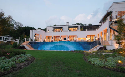 Modern luxury home with illuminated white facade, curved swimming pool, and manicured lawn at dusk
