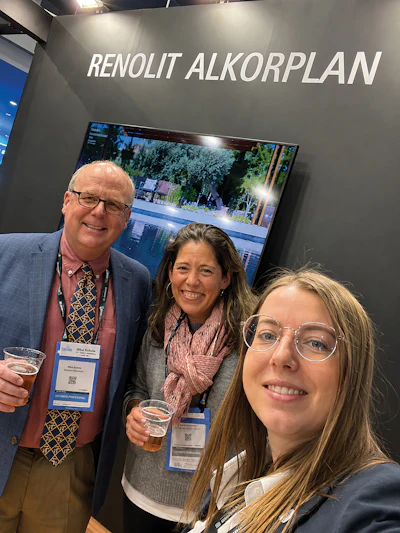Three professionals smiling at trade show booth with Renolit Alkorplan branding and display monitor