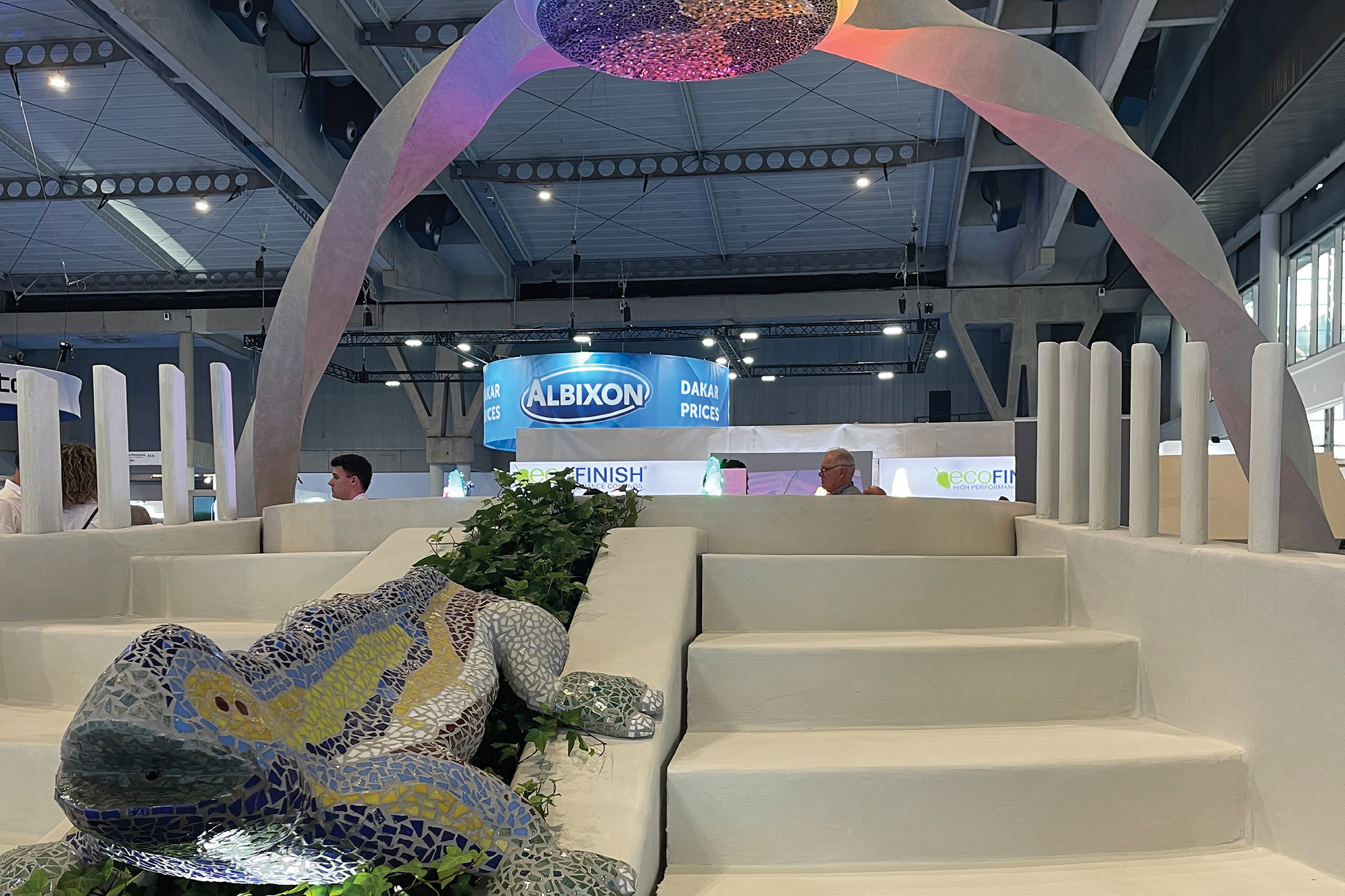 Modern exhibition space with cream-colored architectural elements, colorful overhead installation, and sculptural lizard figure with green foliage