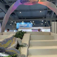 Modern exhibition space with cream-colored architectural elements, colorful overhead installation, and sculptural lizard figure with green foliage