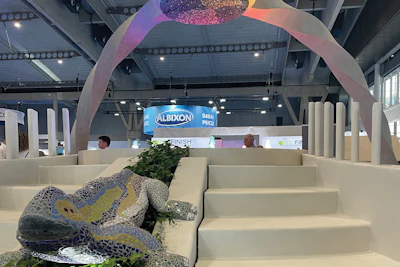 Modern exhibition space with cream-colored architectural elements, colorful overhead installation, and sculptural lizard figure with green foliage