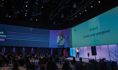 Speaker presenting at corporate conference with turquoise and blue stage backdrop displaying motivational text to seated audience