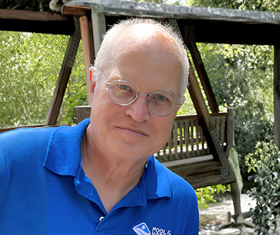Senior man wearing blue polo shirt with logo, smiling at camera outdoors with ivy-covered pergola in background