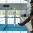 Man in brown jacket standing at indoor swimming pool edge with lane dividers visible