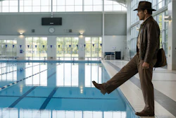 Man in brown jacket standing at indoor swimming pool edge with lane dividers visible