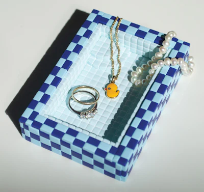 Blue and white checkered jewelry box displaying silver rings, pearl bracelet, and gold necklace with yellow pendant charm
