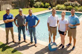 Six team members in casual work attire standing together by a residential swimming pool on a sunny day