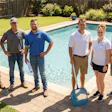 Six team members in casual work attire standing together by a residential swimming pool on a sunny day