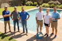 Six team members in casual work attire standing together by a residential swimming pool on a sunny day