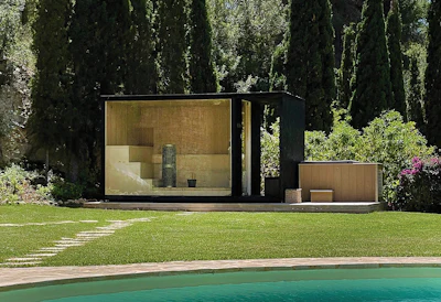 Modern black and wood pool house with glass windows overlooking a turquoise pool in a manicured garden with tall cypress trees
