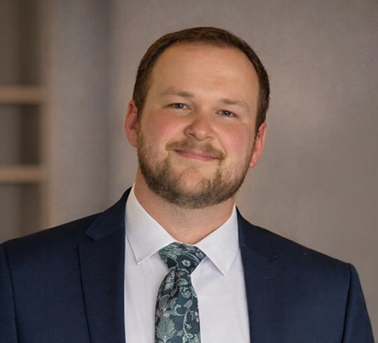 Professional headshot of a man in navy blazer and patterned tie smiling at camera