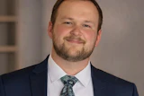 Professional headshot of a man in navy blazer and patterned tie smiling at camera