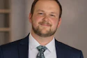Professional headshot of a man in navy blazer and patterned tie smiling at camera