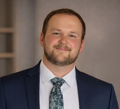 Professional headshot of a man in navy blazer and patterned tie smiling at camera