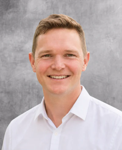Professional headshot of smiling man in white button-up shirt against gray background