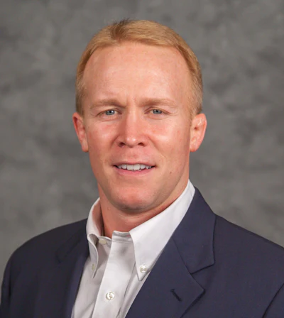 Professional headshot of a man in dark blazer and light shirt smiling against gray background