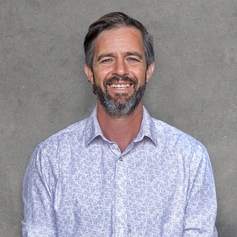 Professional headshot of a smiling man with beard wearing a light blue patterned button-up shirt against a gray background