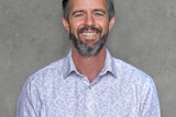 Professional headshot of a smiling man with beard wearing a light blue patterned button-up shirt against a gray background