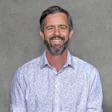 Professional headshot of a smiling man with beard wearing a light blue patterned button-up shirt against a gray background