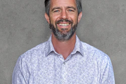 Professional headshot of a smiling man with beard wearing a light blue patterned button-up shirt against a gray background