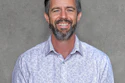 Professional headshot of a smiling man with beard wearing a light blue patterned button-up shirt against a gray background