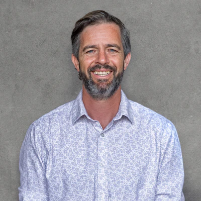 Professional headshot of a smiling man with beard wearing a light blue patterned button-up shirt against a gray background