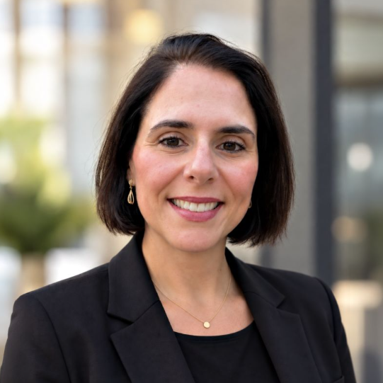 Professional headshot of a woman with dark hair wearing a black blazer, smiling at the camera in an outdoor urban setting
