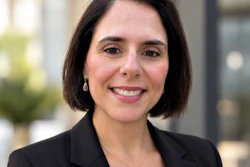 Professional headshot of a woman with dark hair wearing a black blazer, smiling at the camera in an outdoor urban setting