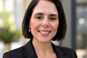 Professional headshot of a woman with dark hair wearing a black blazer, smiling at the camera in an outdoor urban setting