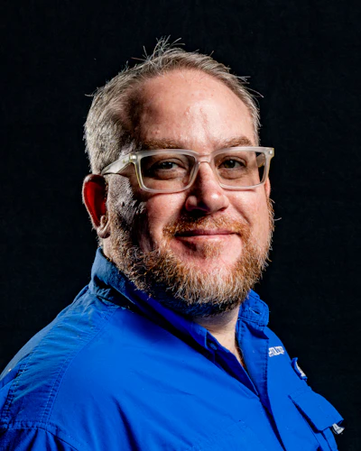 Professional headshot of a bearded man wearing glasses and a blue work shirt against a dark background