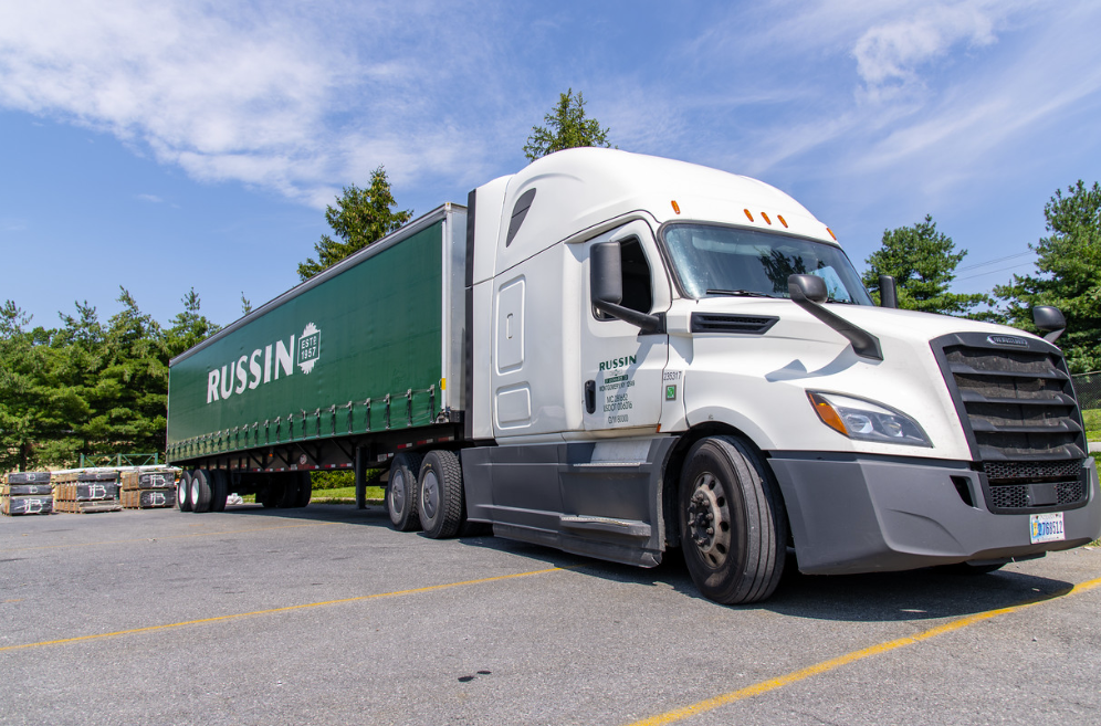 White Freightliner semi-truck with green Russin branded trailer parked in lot with trees