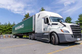 White Freightliner semi-truck with green Russin branded trailer parked in lot with trees