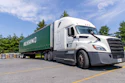 White Freightliner semi-truck with green Russin branded trailer parked in lot with trees