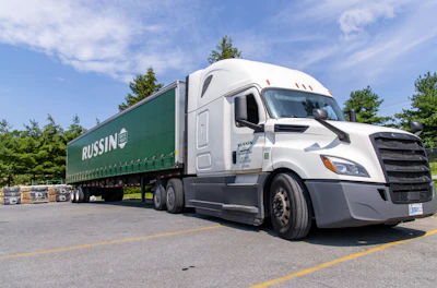 White Freightliner semi-truck with green Russin branded trailer parked in lot with trees