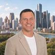 Professional headshot of a man in beige blazer smiling against Detroit skyline backdrop