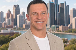 Professional headshot of a man in beige blazer smiling against Detroit skyline backdrop