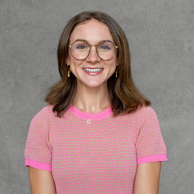Young woman with shoulder-length brown hair wearing clear-framed glasses, pink textured sweater, and gold hoop earrings, smiling at camera against gray background