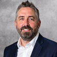 Professional headshot of a bearded man in navy blazer and white shirt smiling against gray background