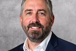 Professional headshot of a bearded man in navy blazer and white shirt smiling against gray background