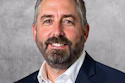 Professional headshot of a bearded man in navy blazer and white shirt smiling against gray background