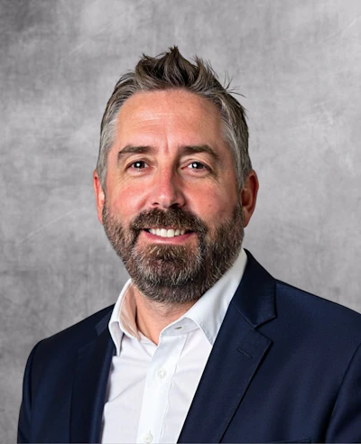 Professional headshot of a bearded man in navy blazer and white shirt smiling against gray background