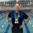 Smiling man wearing three medals standing in front of indoor swimming pool
