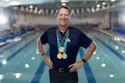 Smiling man wearing three medals standing in front of indoor swimming pool